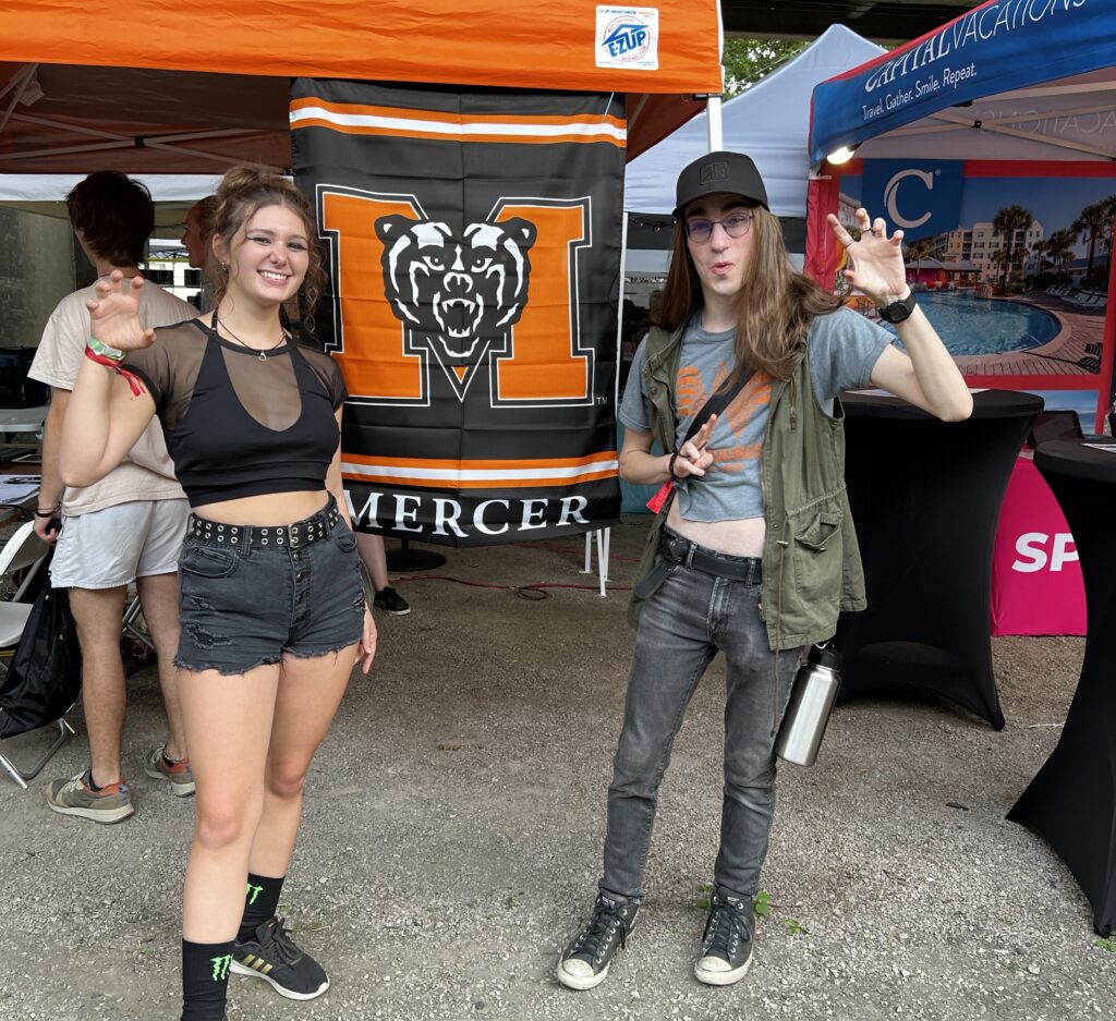 Two people pose playfully in front of a Mercer banner at an outdoor event. Both have their hands raised like claws.