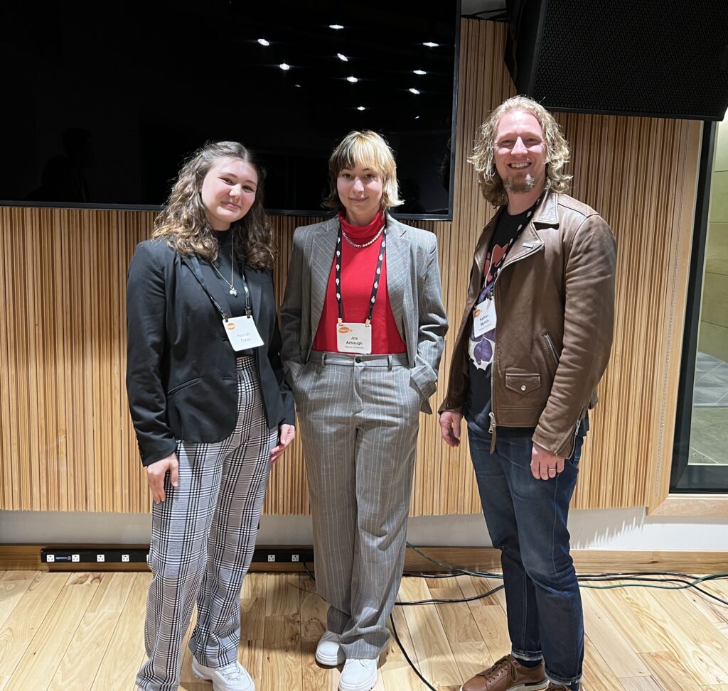 Three people standing together indoors, dressed in business casual attire, smiling at the camera, with a wood-paneled wall behind them.