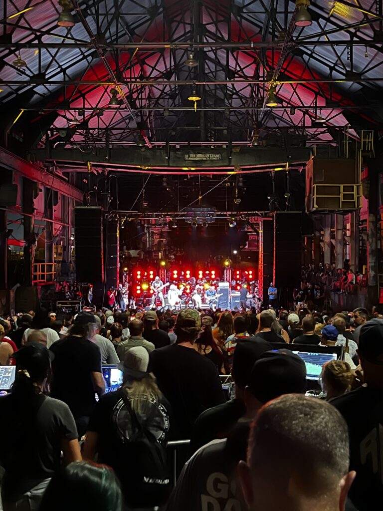 A crowd watches a band perform on stage in an industrial-style venue with overhead metal beams and red lighting.