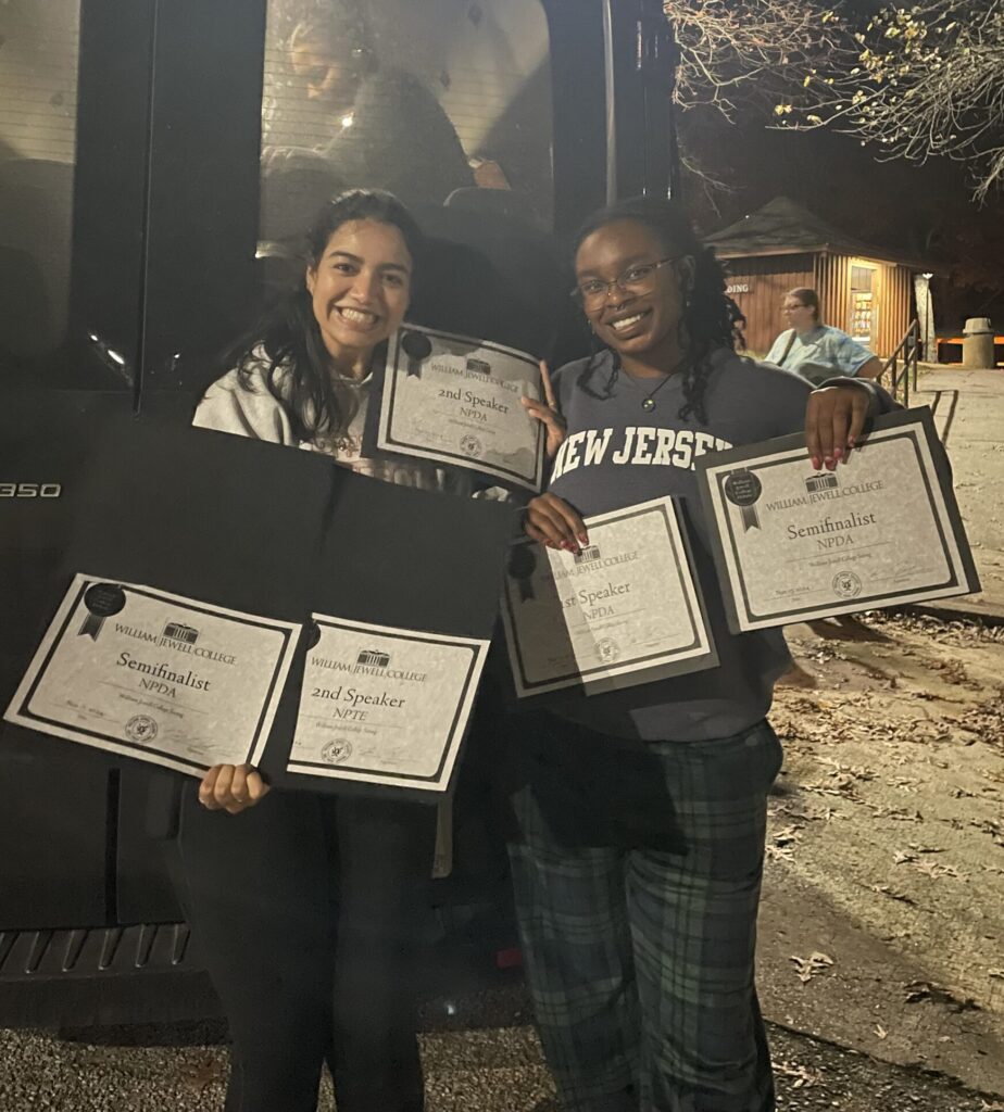 Two people are standing outside holding four certificates, smiling. A person is visible inside the vehicle behind them.