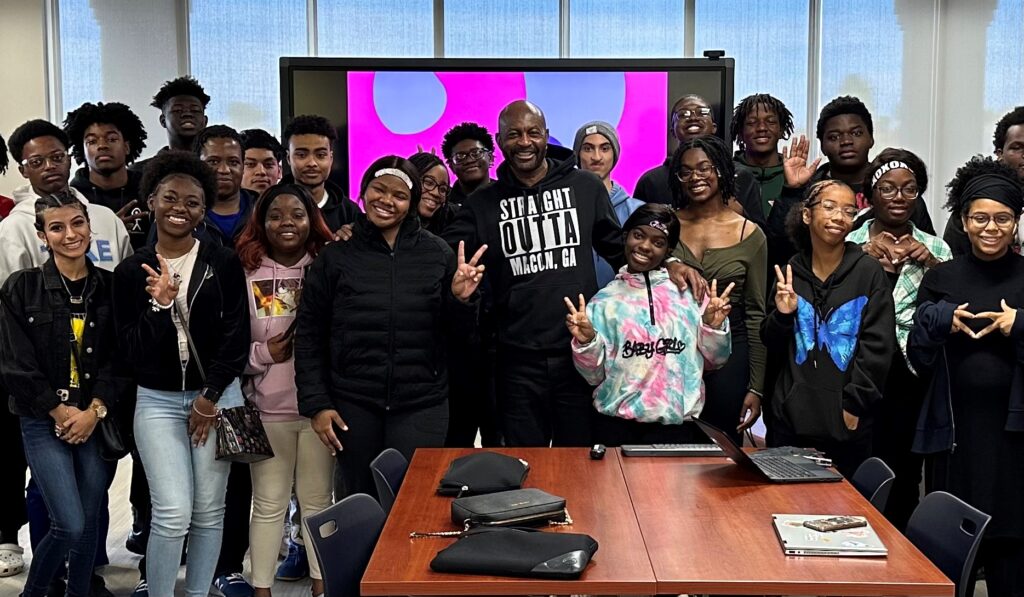 A group of students and an adult gather in a classroom, smiling and posing in front of a screen displaying a colorful image.