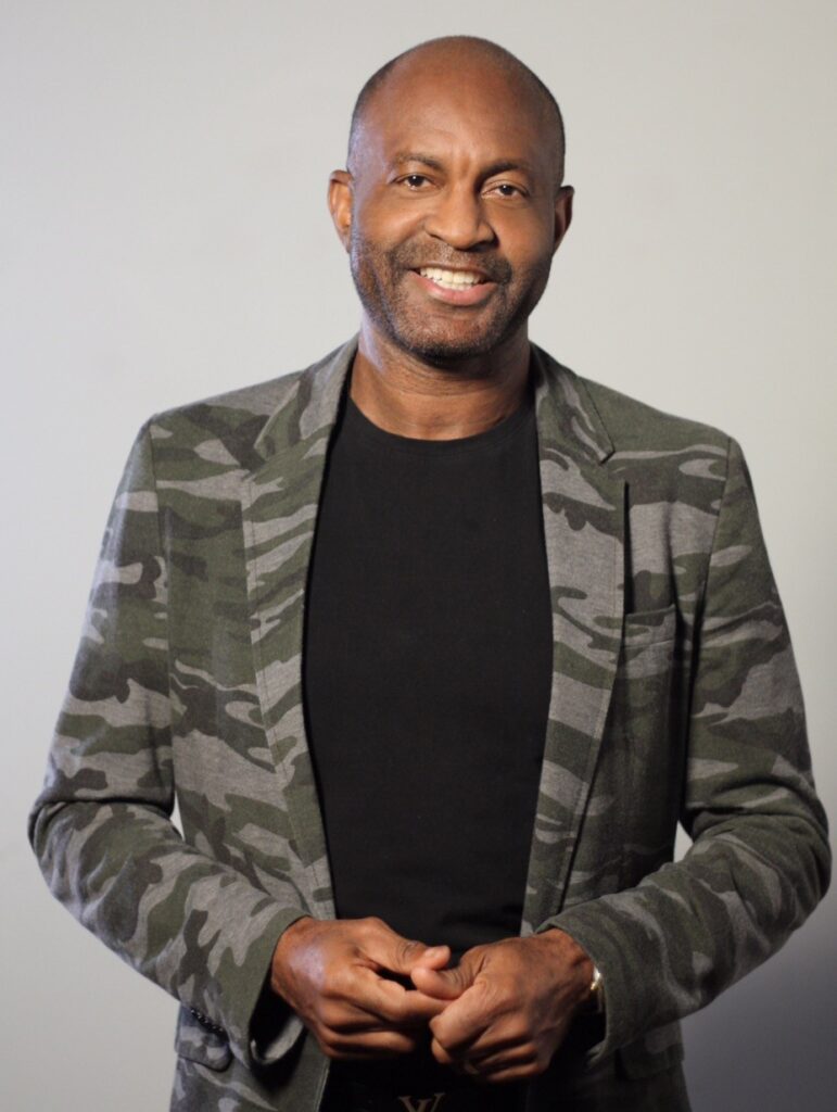 A person wearing a camo blazer and black shirt smiles at the camera.