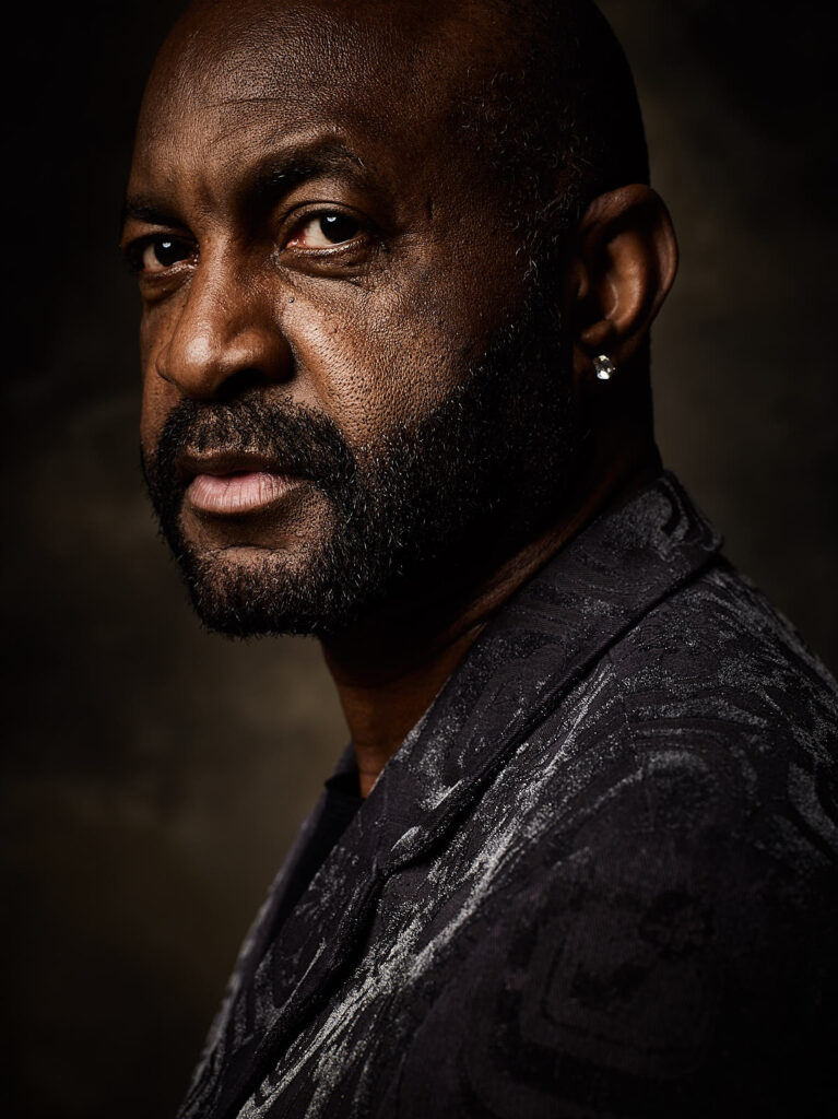 A man with a beard and an earring wearing a patterned jacket looks directly at the camera against a dark background.