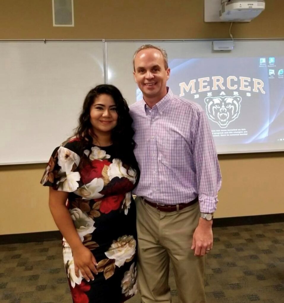 Mary Ann Santos and Jason Harper stand in a classroom in front of a whiteboard and a projector screen displaying Mercer Bears.