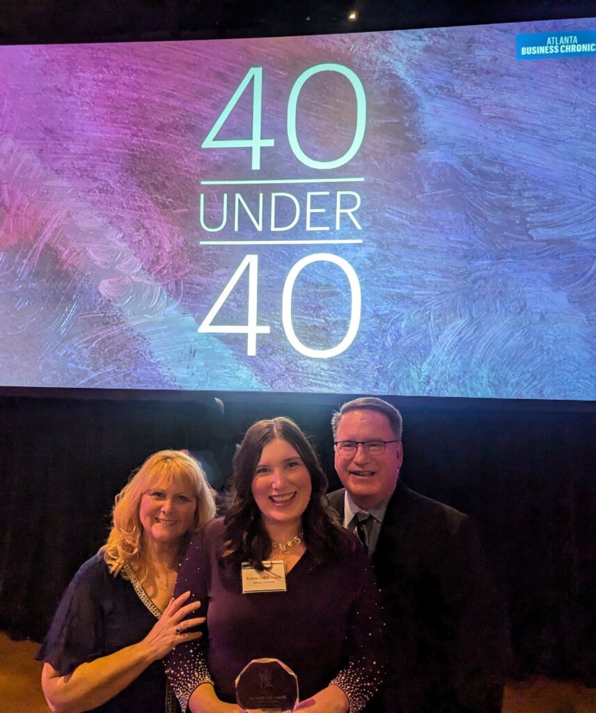 Three people pose with an award in front of a 40 Under 40 sign at an event.