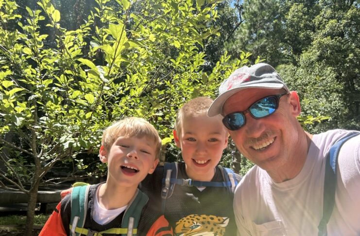 Dr. Craig McMahan and two 9-year-old boys smile for a selfie outdoors, with trees and sunlight in the background.