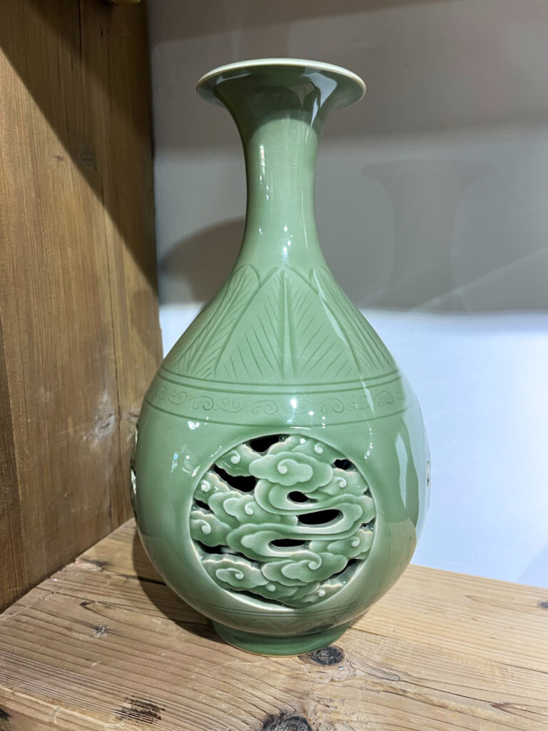 Green ceramic vase with intricate carved designs, featuring a tall neck and placed on a wooden surface.