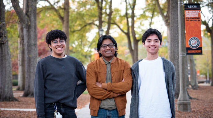 Mercer freshmen create video game for technical communication class Three people stand smiling in a park with trees and a campus banner in the background.