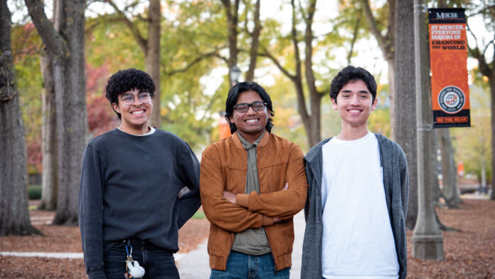 Three people stand smiling in a park with trees and a campus banner in the background.