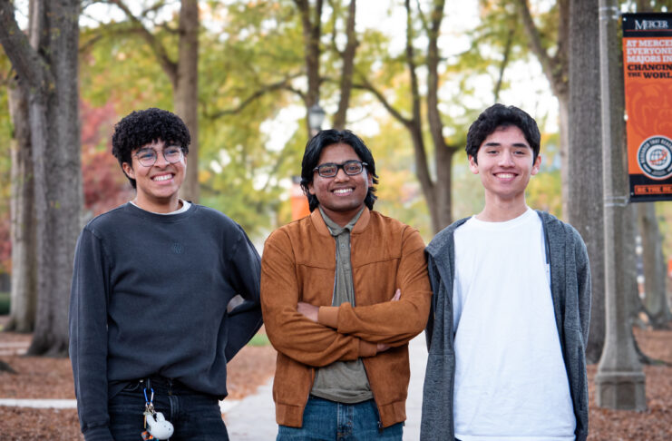 Three people stand smiling in a park with trees and a campus banner in the background.