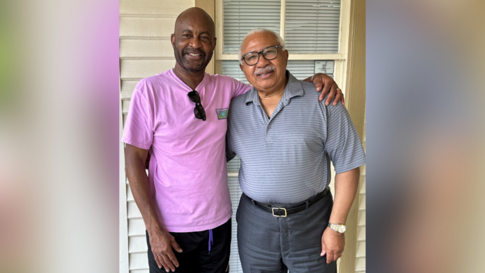 Two men standing together, smiling, with one wearing a pink shirt and the other in a gray polo.