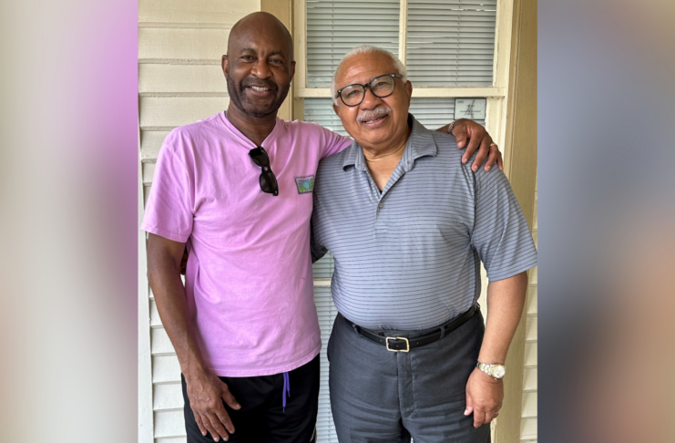 Two men standing together, smiling, with one wearing a pink shirt and the other in a gray polo.