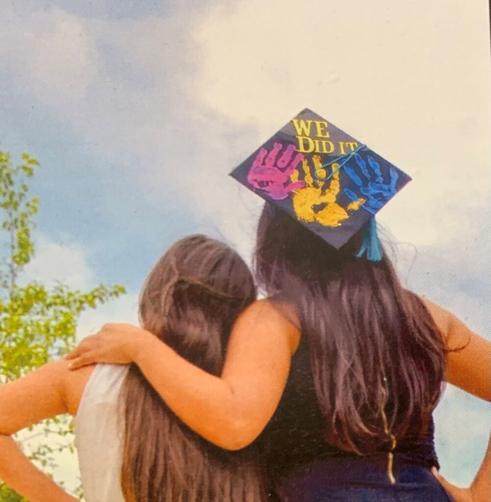 Two graduates embrace outdoors, one wearing a cap with colorful handprints and the words We Did It! against a cloudy sky.