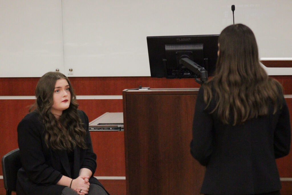 Two women face each other in a courtroom setting. Kasey Hoyt is sitting and the one standing.