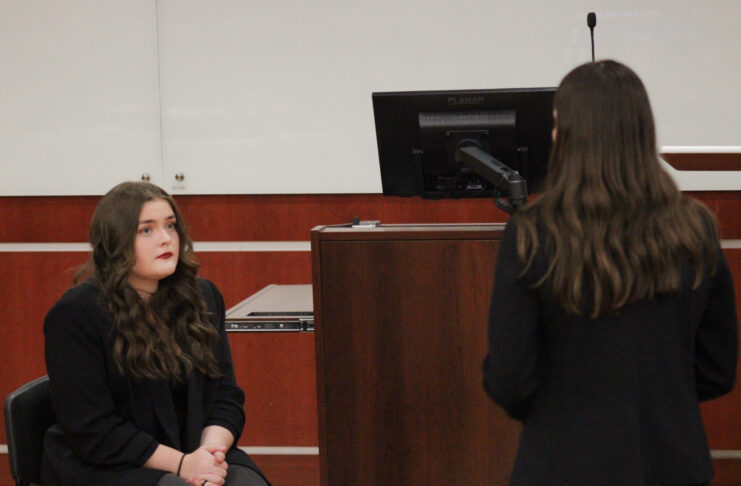 Two women face each other in a courtroom setting. Kasey Hoyt is sitting and the one standing.