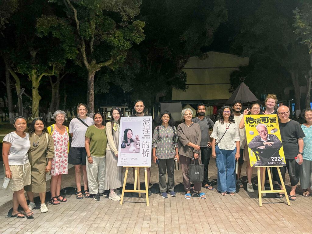 A group of people stand outdoors at night beside two posters, surrounded by trees.