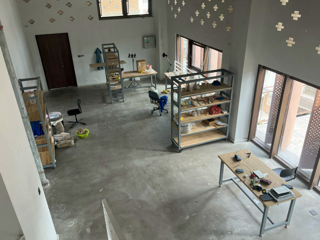 Aerial view of a spacious room with shelves, tables, chairs, and various items, including ceramics and tools, on concrete flooring.