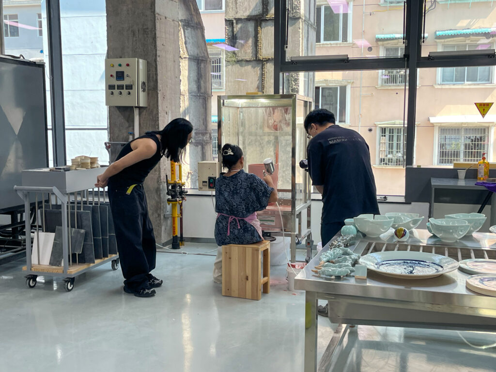 Three people are working in a ceramic studio, surrounded by pottery and equipment, near large windows.