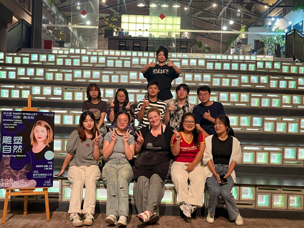 Group of eleven people sitting and standing on steps indoors, with a poster to the left side.