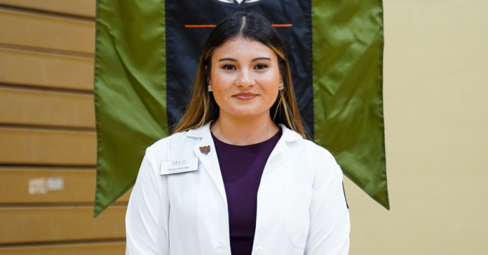 A person in a white coat stands indoors, wearing a name badge, in front of a flag with wooden paneling to the left.