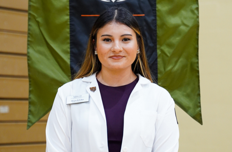 A person in a white coat stands indoors, wearing a name badge, in front of a flag with wooden paneling to the left.