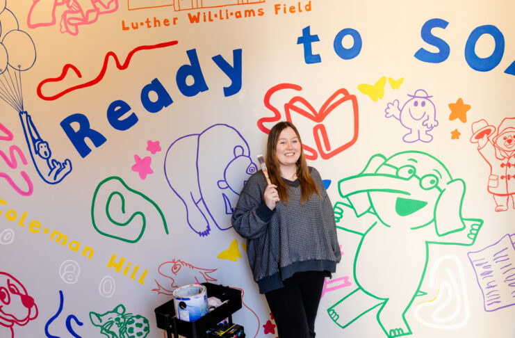 Abby Milam standing in front of a colorful mural with cartoon illustrations and the text Ready to SOAR on a wall.