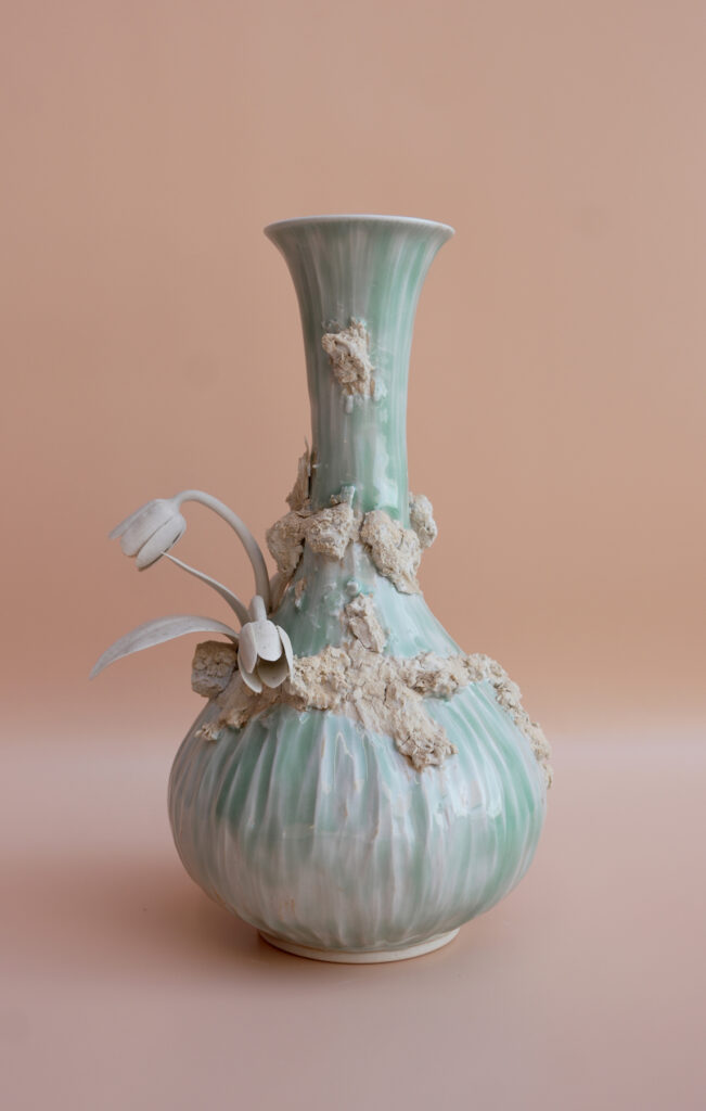 A green ceramic vase with a tall neck, adorned with rough textures and two white sculptural flowers on a peach background.