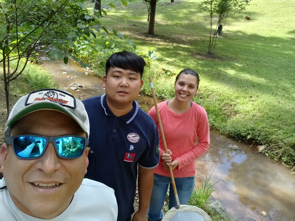 Three people outdoors by a creek, holding nets and smiling; two standing and one wearing sunglasses in the foreground.
