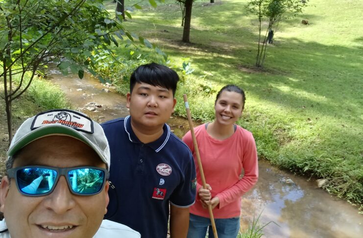 Three people outdoors by a creek, holding nets and smiling; two standing and one wearing sunglasses in the foreground.