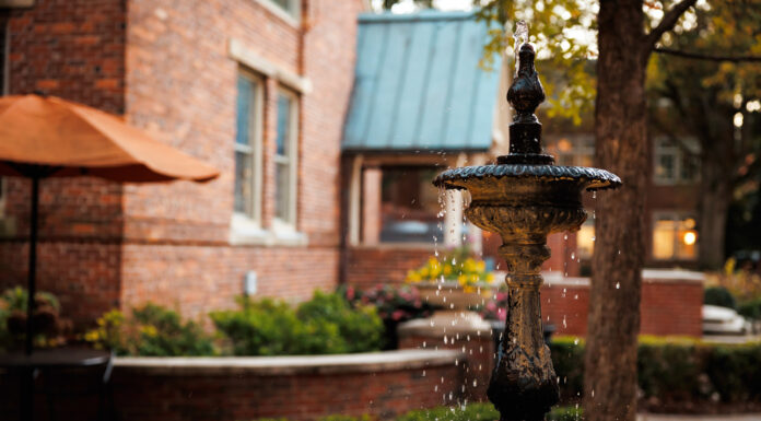 Faculty and Staff Notables | February 2025 A decorative black metal fountain with flowing water in a garden courtyard, surrounded by brick buildings and greenery.
