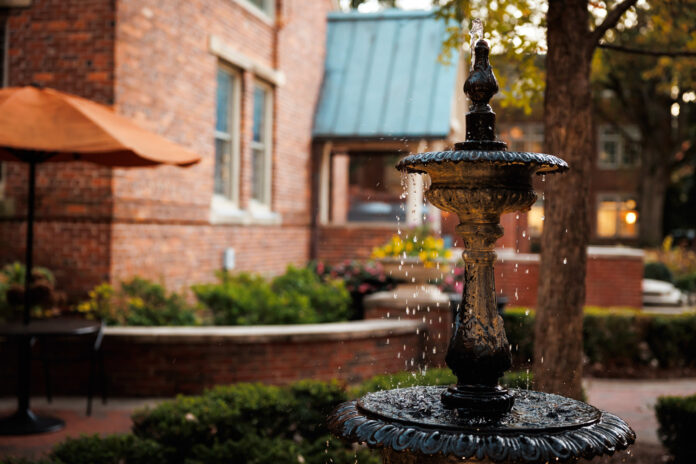 A decorative black metal fountain with flowing water in a garden courtyard, surrounded by brick buildings and greenery.