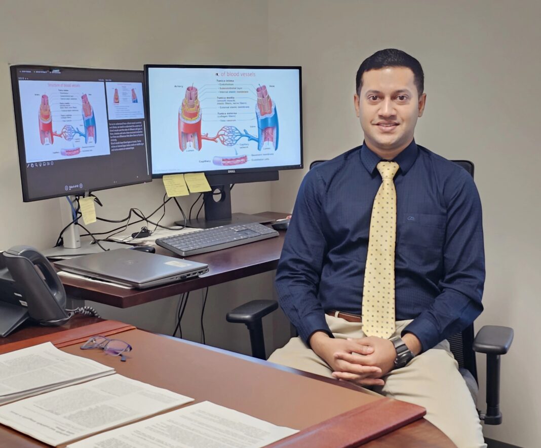 Dr. Raquib Hasan in a blue shirt and yellow tie sits at a desk with two monitors displaying medical diagrams and articles.