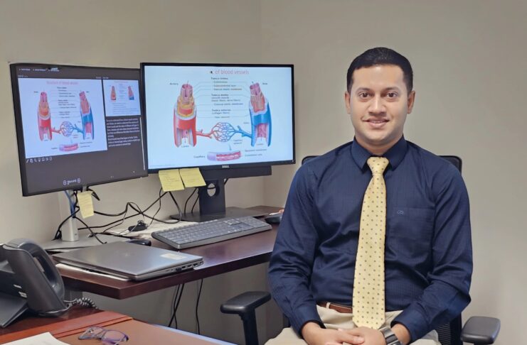 Dr. Raquib Hasan in a blue shirt and yellow tie sits at a desk with two monitors displaying medical diagrams and articles.