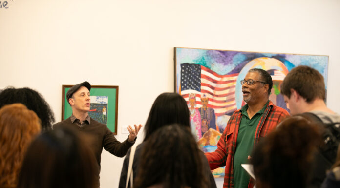 Ads for runaway slaves tell story of Macon’s past in student-curated exhibit Two men stand in front of an audience, speaking animatedly, with colorful artwork featuring an American flag in the background.
