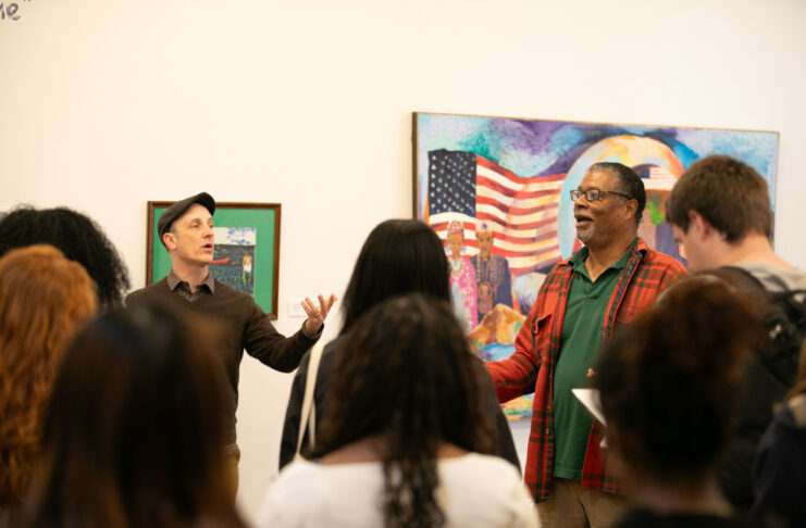 Two men stand in front of an audience, speaking animatedly, with colorful artwork featuring an American flag in the background.