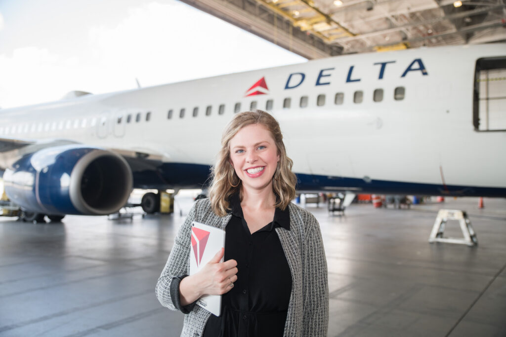 A person stands smiling in front of a Delta airplane inside a hangar, holding a notebook with a matching logo.