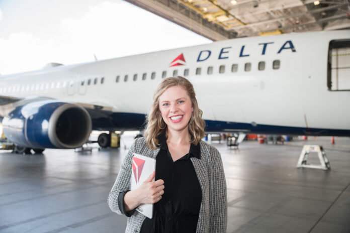 A person stands smiling in front of a Delta airplane inside a hangar, holding a notebook with a matching logo.