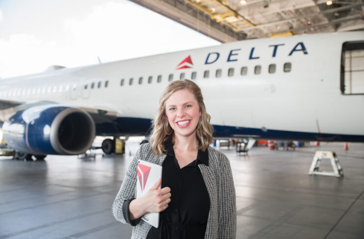 A person stands smiling in front of a Delta airplane inside a hangar, holding a notebook with a matching logo.