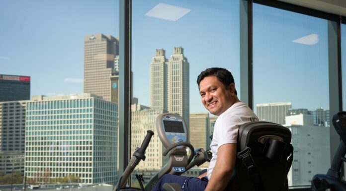 Alumnus accompanies patients on recovery journey as physical therapist Hemal Patel sitting on an exercise bike, smiling with a city skyline visible through large windows in the background.