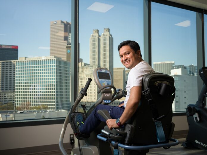 Hemal Patel sitting on an exercise bike, smiling with a city skyline visible through large windows in the background.