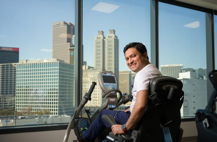 Hemal Patel sitting on an exercise bike, smiling with a city skyline visible through large windows in the background.