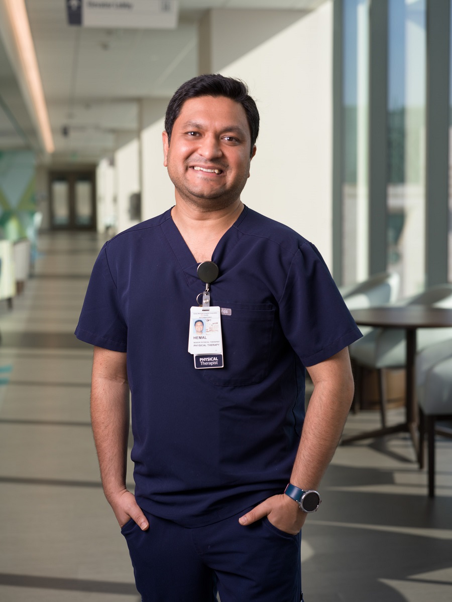 A healthcare professional in navy scrubs stands in a well-lit corridor, wearing a badge and a wristwatch, smiling slightly.