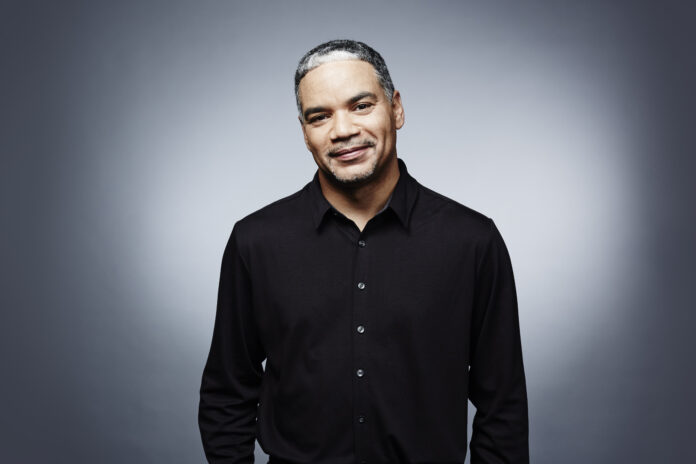 Man in a black shirt standing against a gradient gray background, looking directly at the camera with a slight smile.