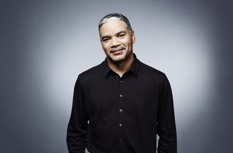 Man in a black shirt standing against a gradient gray background, looking directly at the camera with a slight smile.