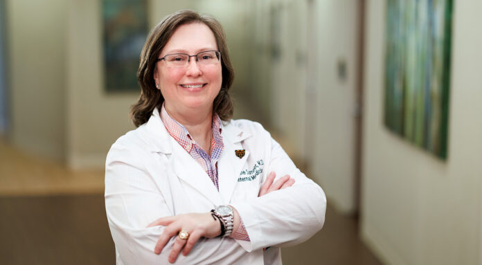 Dr. Loraine Sumner in a white lab coat smiling and standing with folded arms in a hallway.