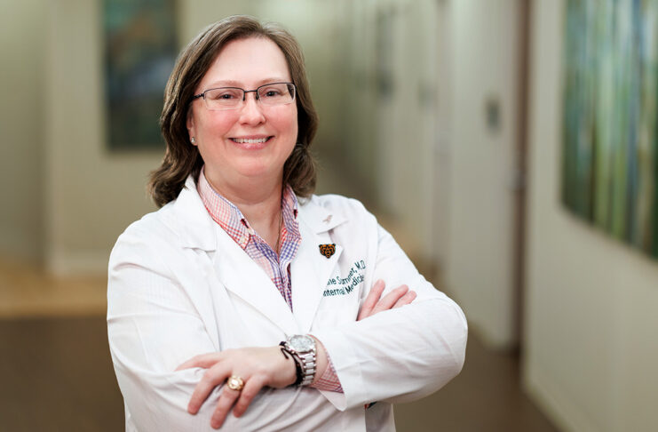 Dr. Loraine Sumner in a white lab coat smiling and standing with folded arms in a hallway.