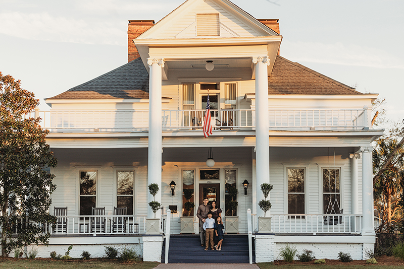 A family of three stands on the porch steps of a large white house with columns and an American flag.