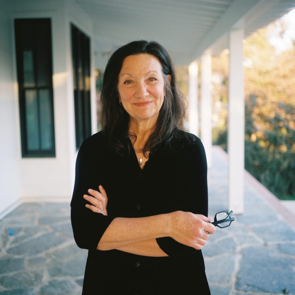 A woman with long dark hair and a black blouse stands on a porch, arms crossed, holding glasses, with a slight smile.
