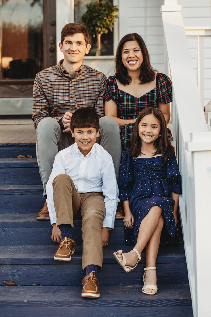 A family of four sits on outdoor stairs; two adults and two children smile at the camera.