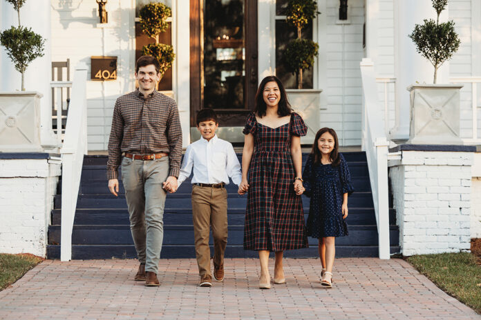 The O'Connor family of four, dressed in casual attire, walks hand in hand on a brick path in front of a white house with blue stairs.